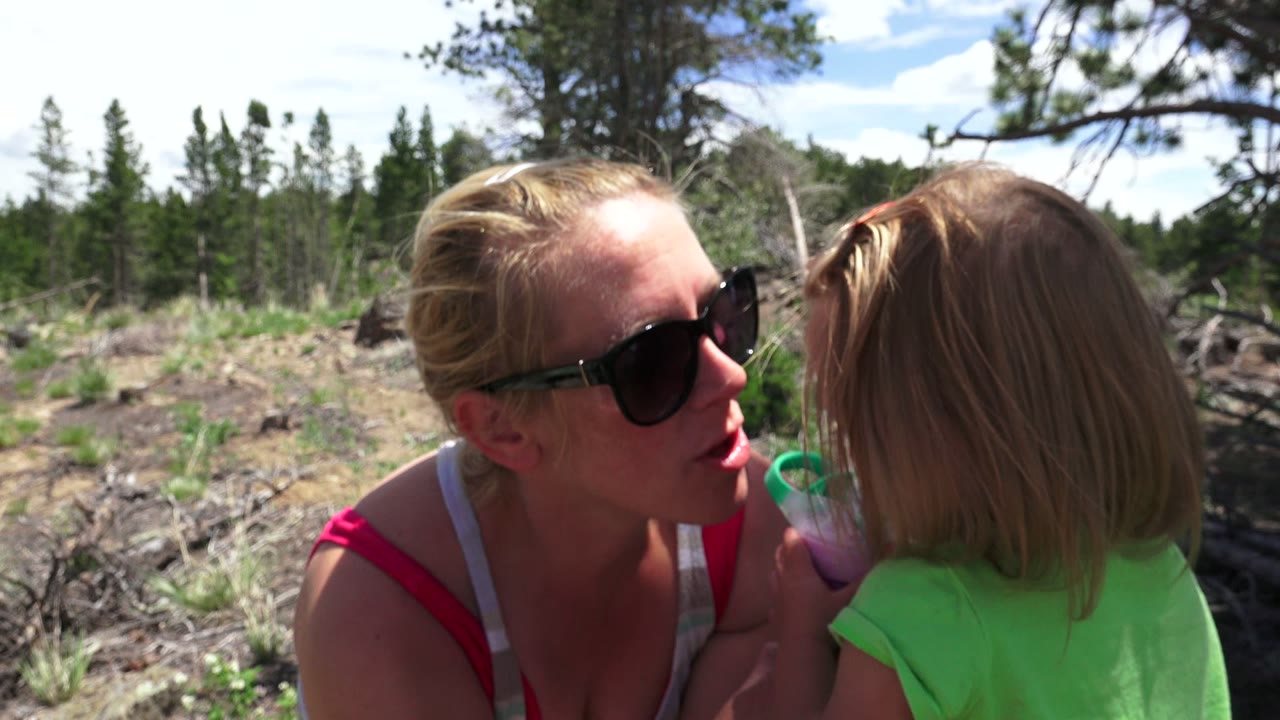 In Westminster, Colorado, the mother leans closer toward her daughter in a face-to-face moment, with the child's cup raised and a rocky wooded area behind them.
