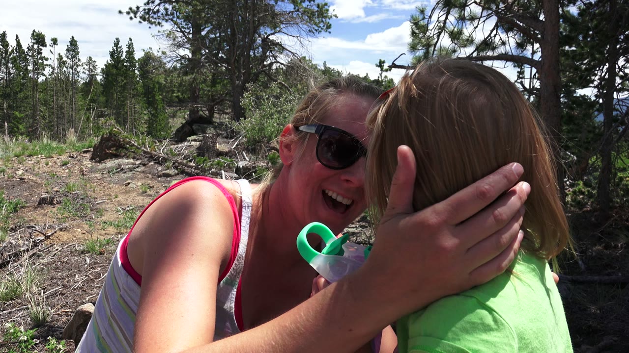 In Westminster, Colorado, an adult woman in sunglasses laughs beside a young girl in a bright green shirt, with trees and rocky ground behind them.