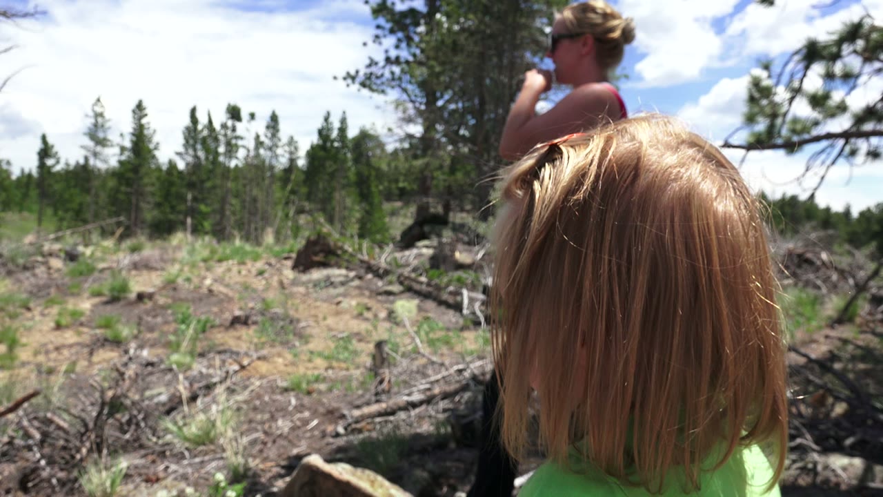At 0:01 in Westminster, Colorado, the child's blond hair remains close to the lens while an adult woman in sunglasses stands in the midground beside scattered branches and low vegetation.