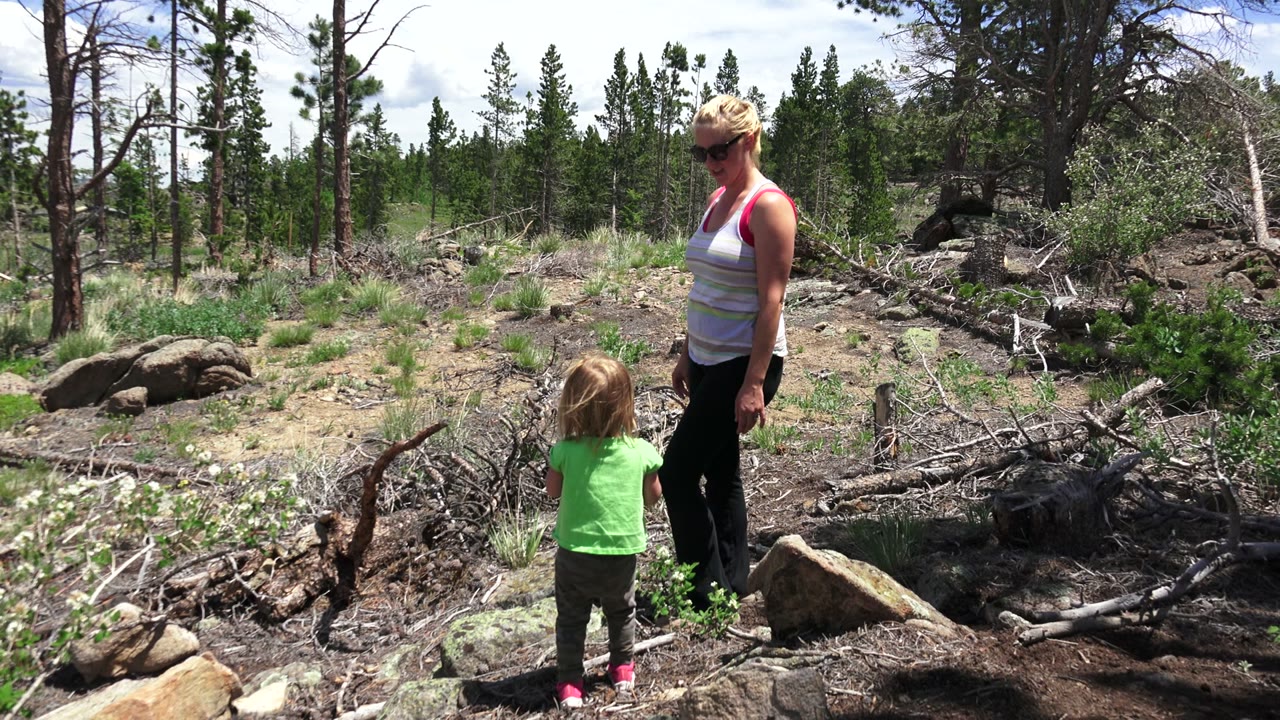At 0:00, a woman in sunglasses stands beside a young child in a green shirt among rocks, dry branches, shrubs, and pine trees in Westminster, Colorado.