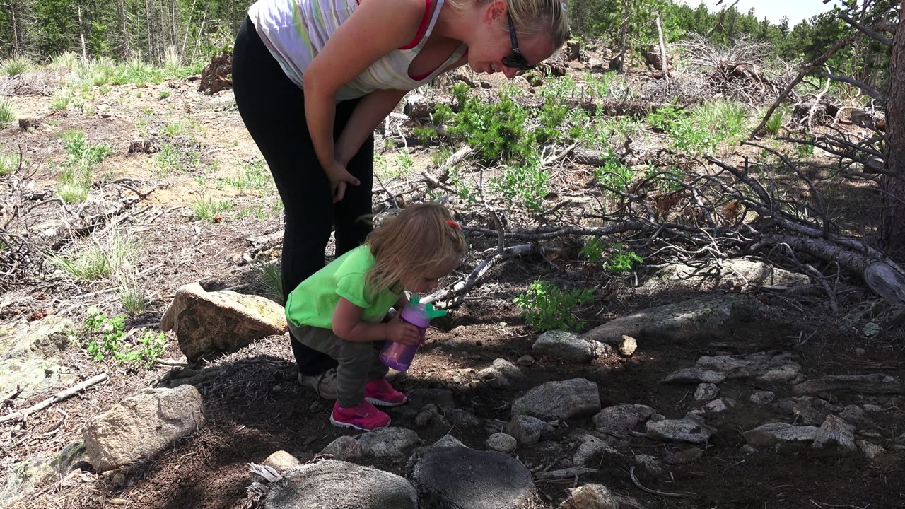 Mother bends over young girl examining rocks on a dirt trail.