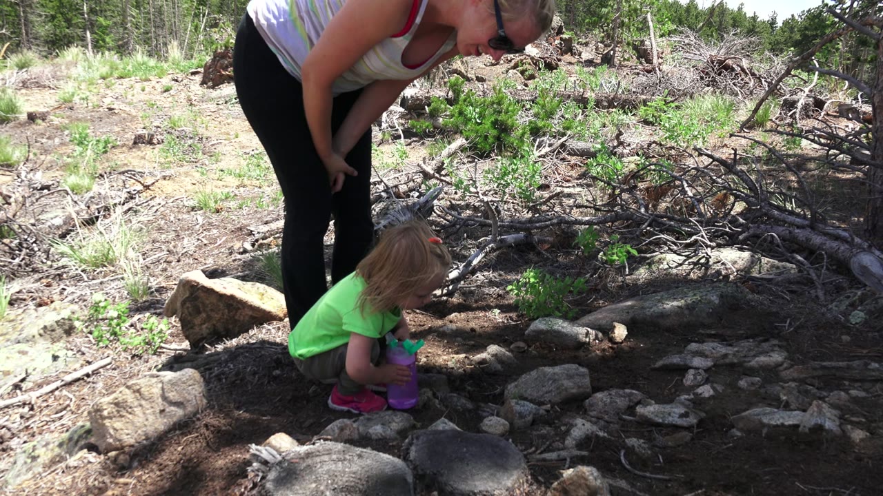 At 0:01 in Westminster, Colorado, the girl crouches lower with a purple cup while her mother watches from above beside scattered stones and fallen branches.