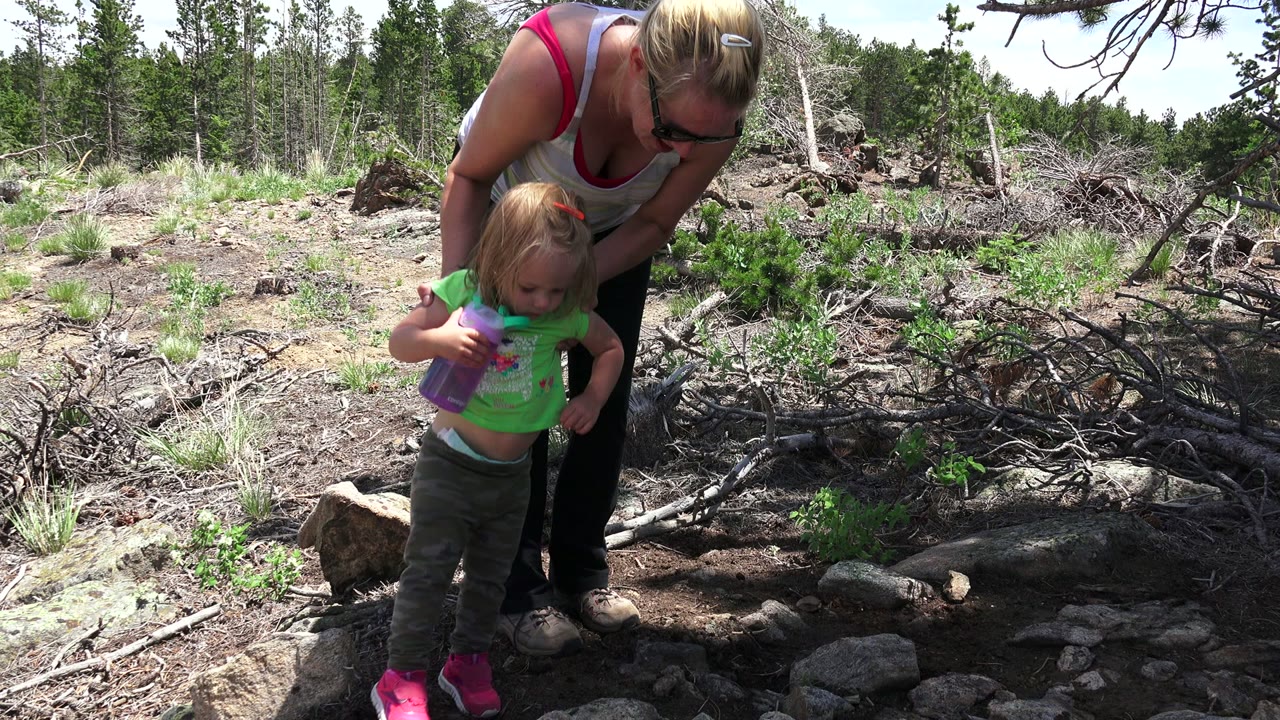 In Westminster, Colorado, the child faces forward with a purple cup as the woman leans close beside her in the sunlit rocky garden.
