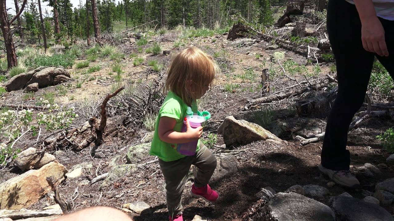 Young girl holding a purple bottle near rocks and a standing adult's legs.