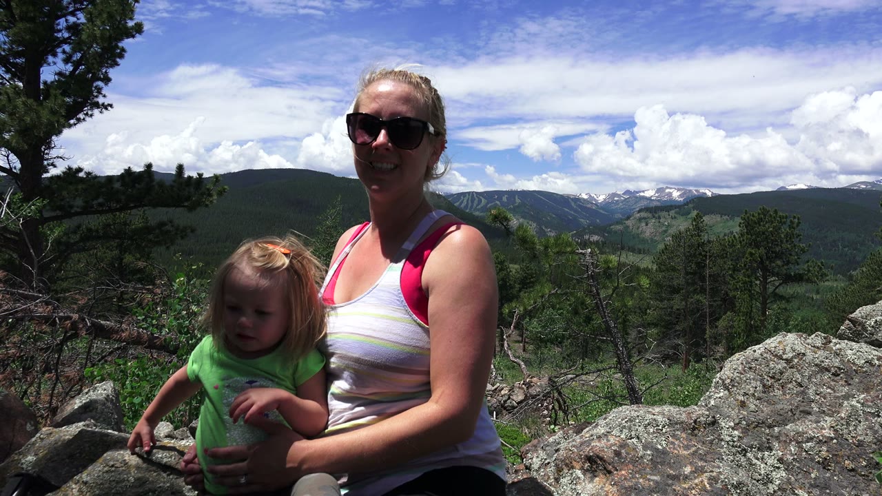 Woman in sunglasses holding a child beside rocks with mountains behind them.