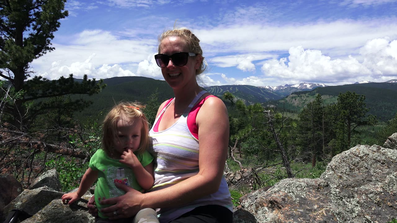 Woman in sunglasses holding a child on rocks with mountains behind them.