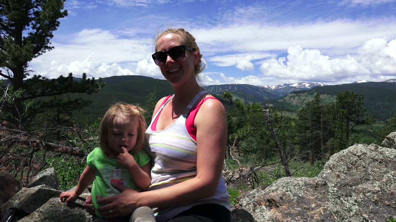 At 0:01, the woman smiles toward the camera while the child sits close to her on the rocks, with trees and layered mountain ridges visible across the background in Westminster, Colorado.