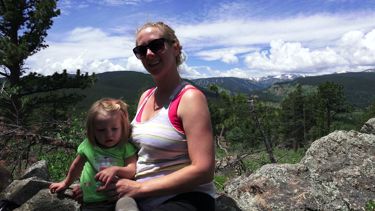 At 0:00, a woman in sunglasses and a small girl sit close together on rocks in Westminster, Colorado, with pine trees and distant green mountains behind them.