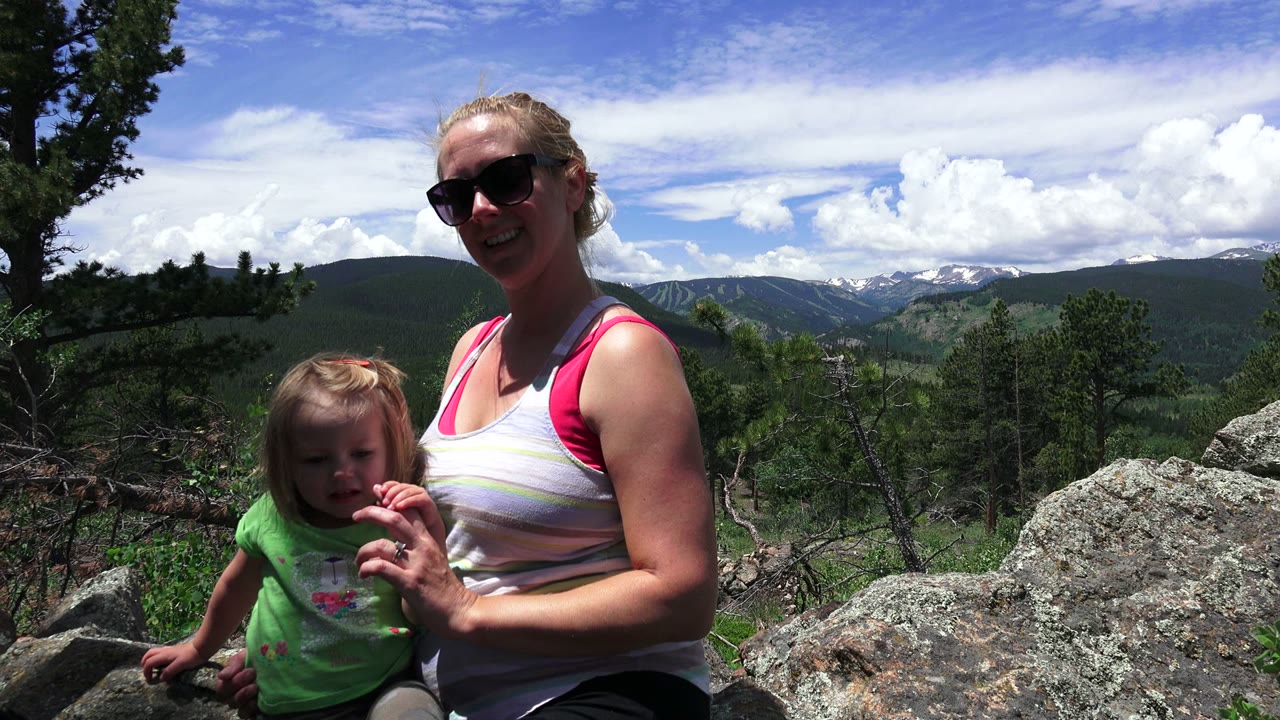 At 0:01, the small girl faces the camera more directly while the woman smiles beside her on rocks in Westminster, Colorado, with a broad mountain backdrop.