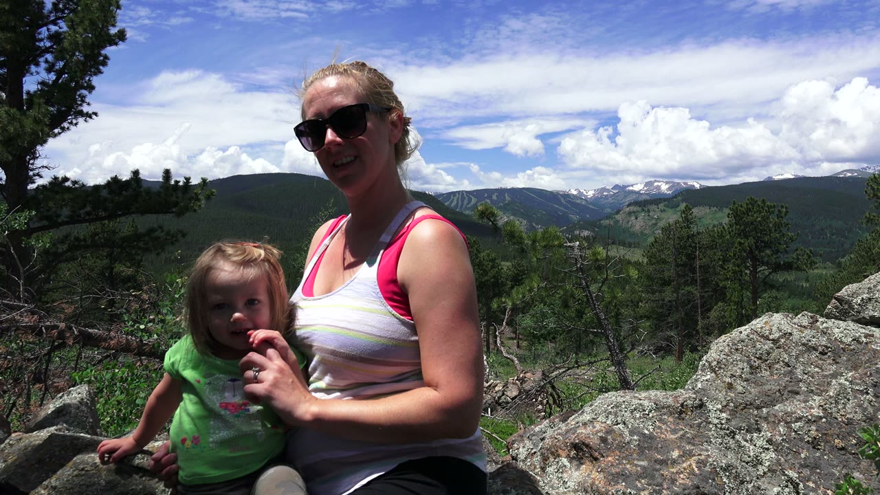 Woman smiles beside a small girl on rocks under bright sky.