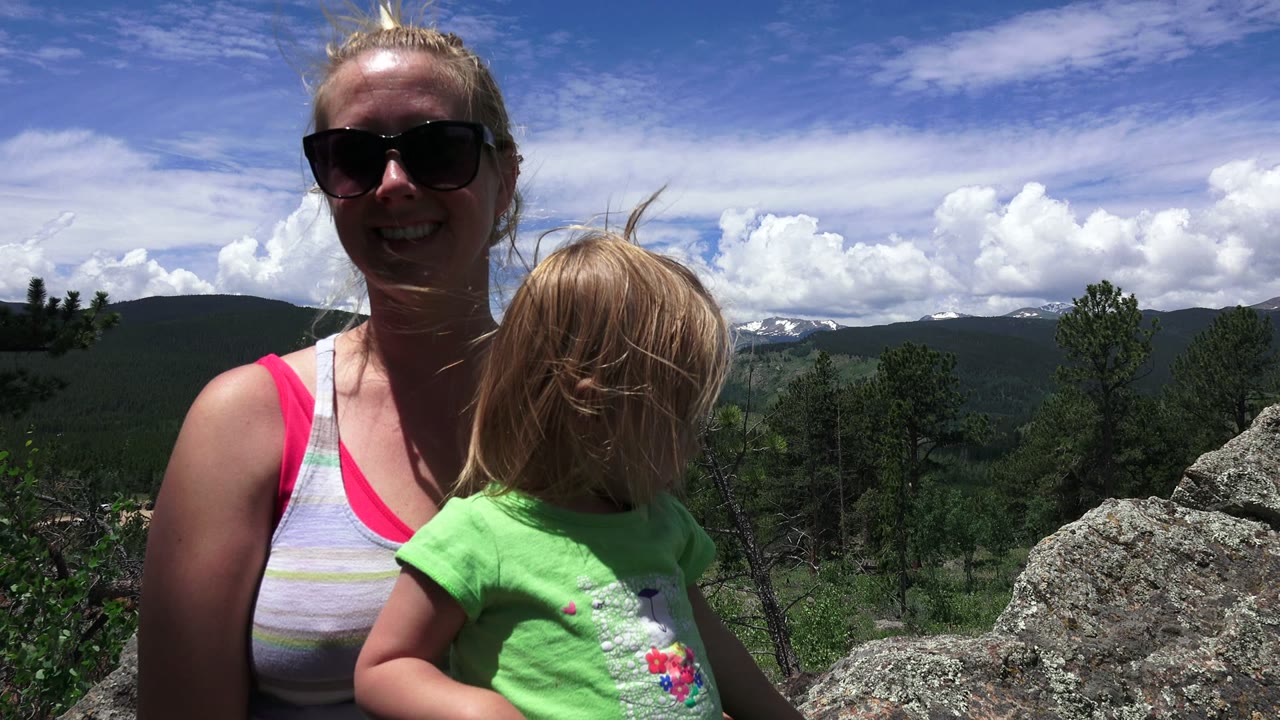 Woman in sunglasses holding a young child at a mountain overlook.