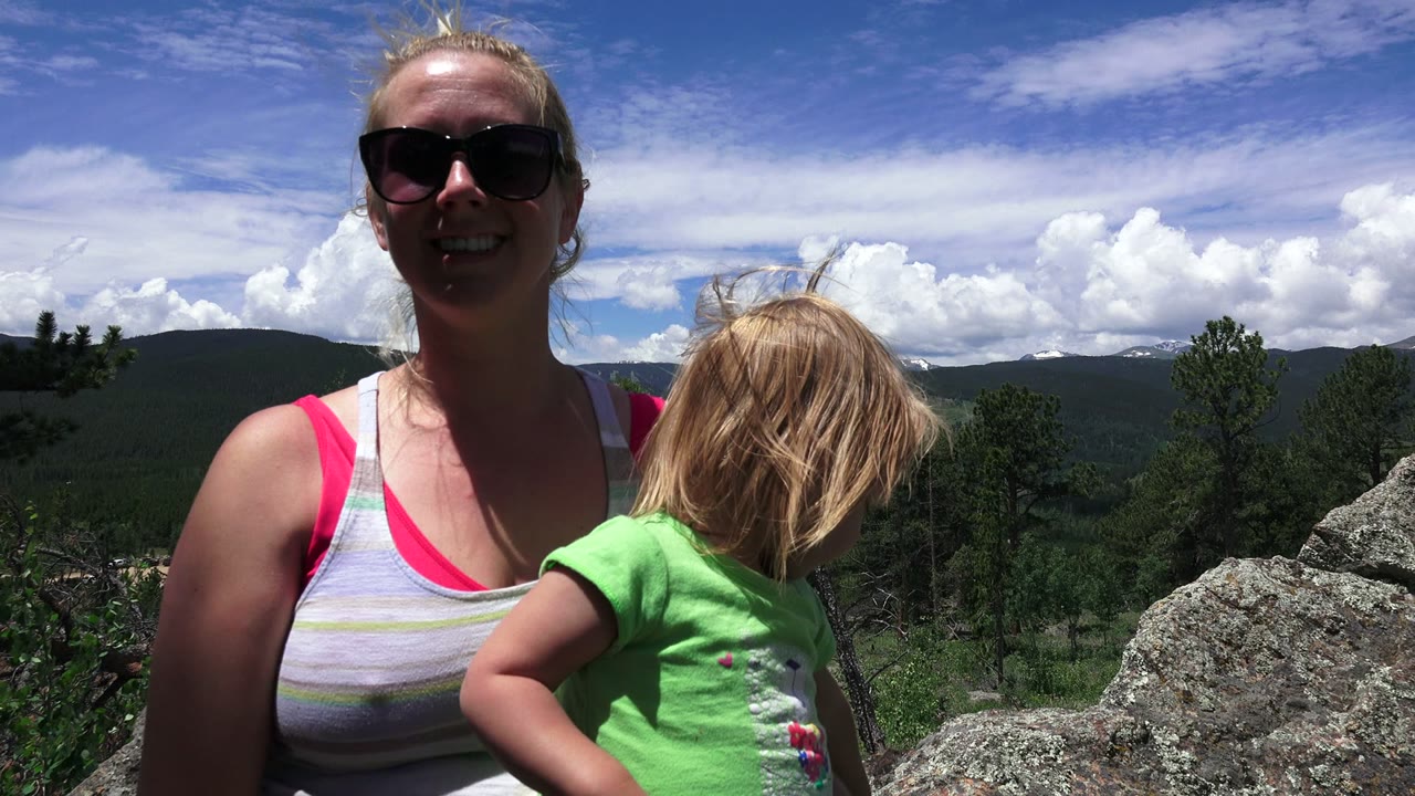 In Westminster, Colorado, the woman faces forward in sunglasses while the child turns downward, with textured rocks and a forested ridge in the background.