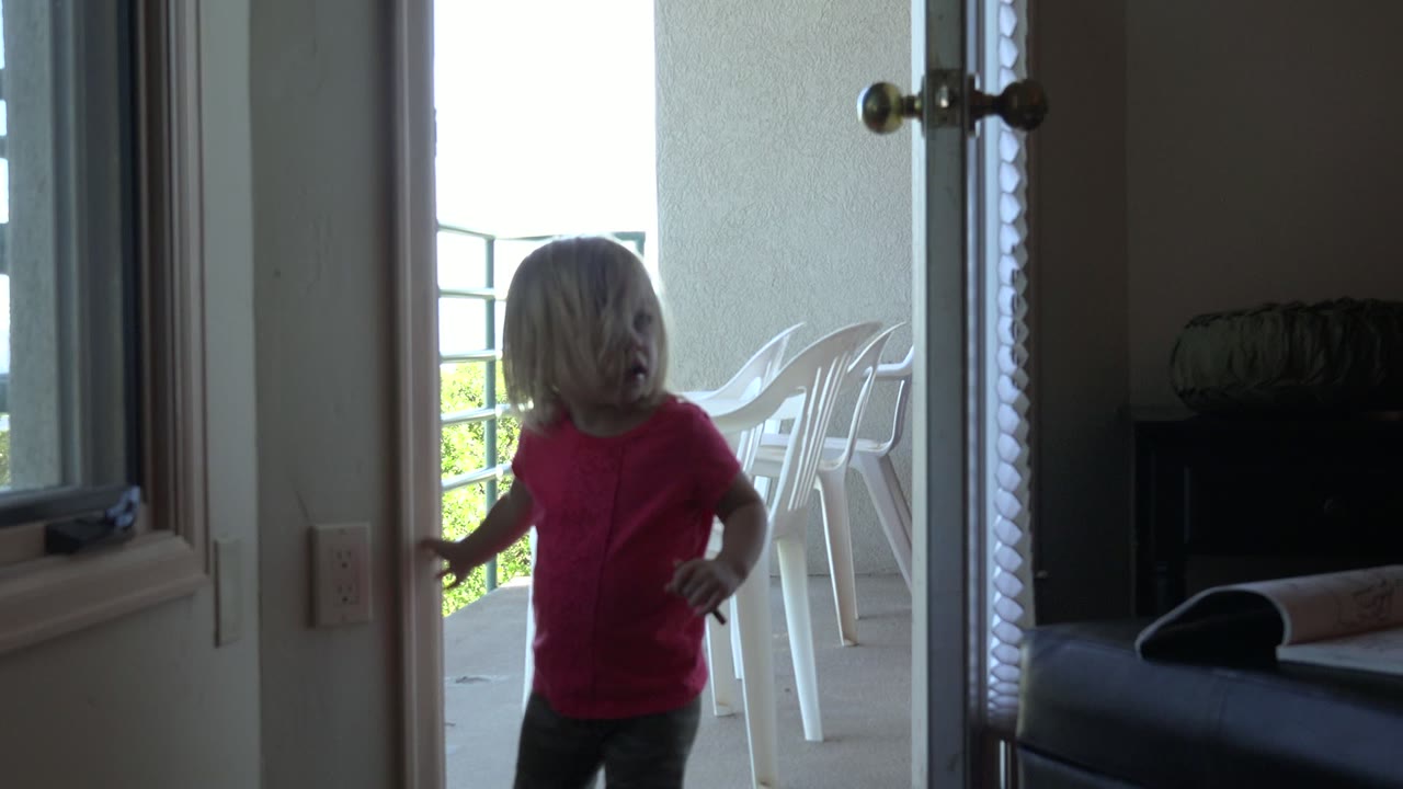 The girl pauses by the door handle in the Mesa, Colorado interior, framed by pale walls, the open door, and white plastic chairs outside.