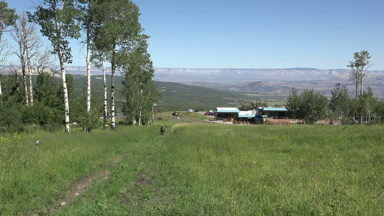 Mesa, Colorado hillside view with a grassy foreground, a thin dirt track, clustered trees, small buildings and vehicles in the distance, and faint tiny people near the field.