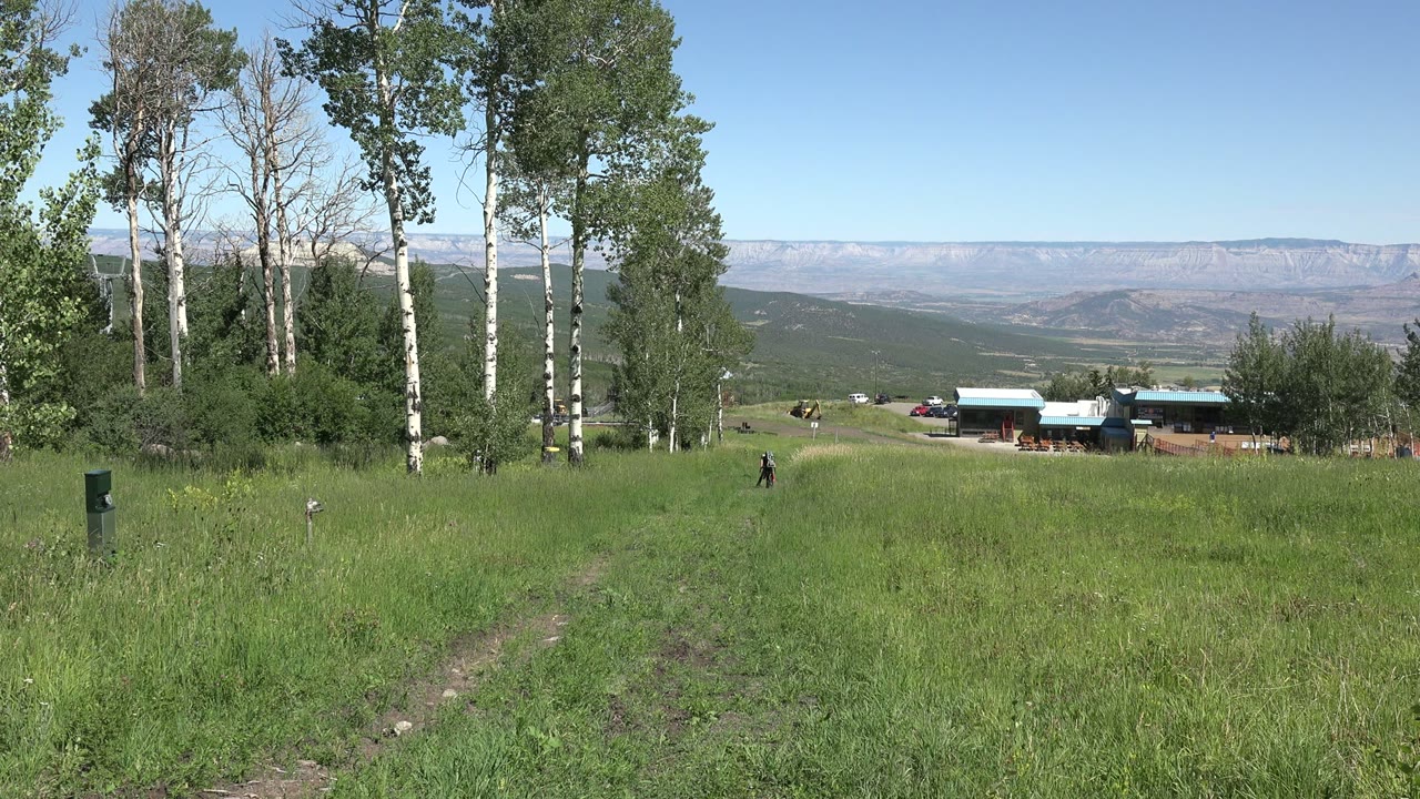 At 0:01, the Mesa, Colorado view holds on a sloping green field, clustered trees, low ranch buildings, scattered horses, and a broad mesa horizon.
