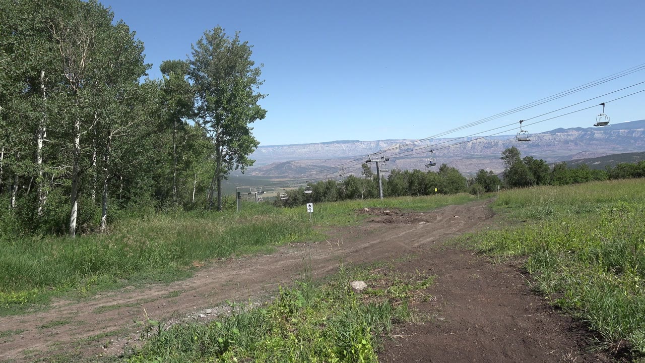 Opening frame of a Mesa, Colorado dirt trail bordered by green grass and tall trees, with cable cars on overhead lines and distant mountains under clear sky, no visible people.