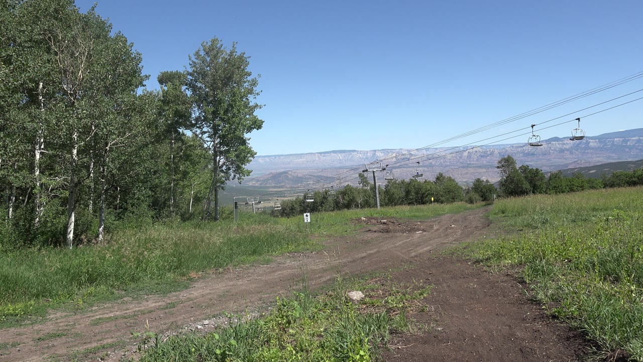 A slightly shifted view of the Mesa, Colorado trail shows more open dirt in the foreground, dense grass at right, trees at left, and cable cars suspended against the blue sky, no visible people.