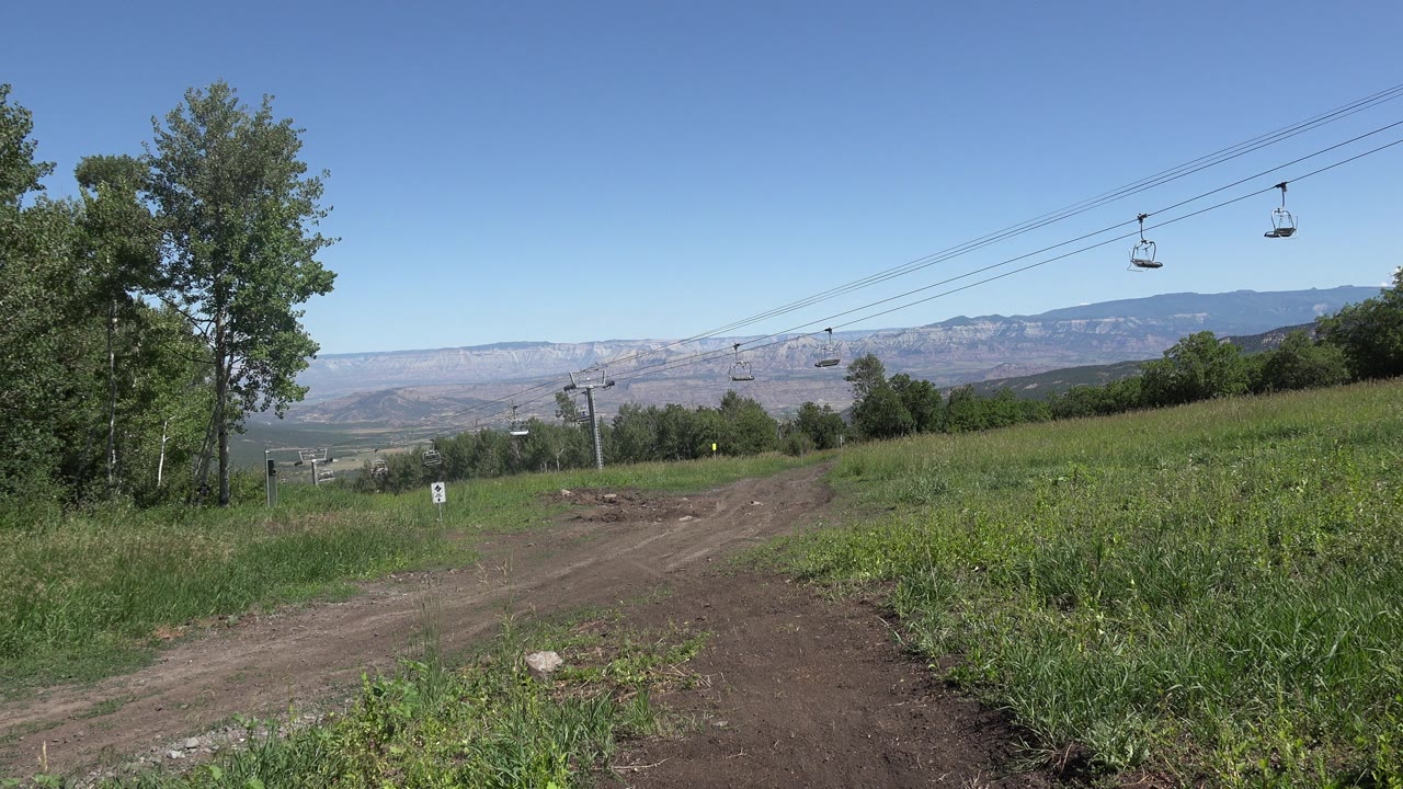 Dirt path through green grass with chairlift cables overhead and mountains beyond.