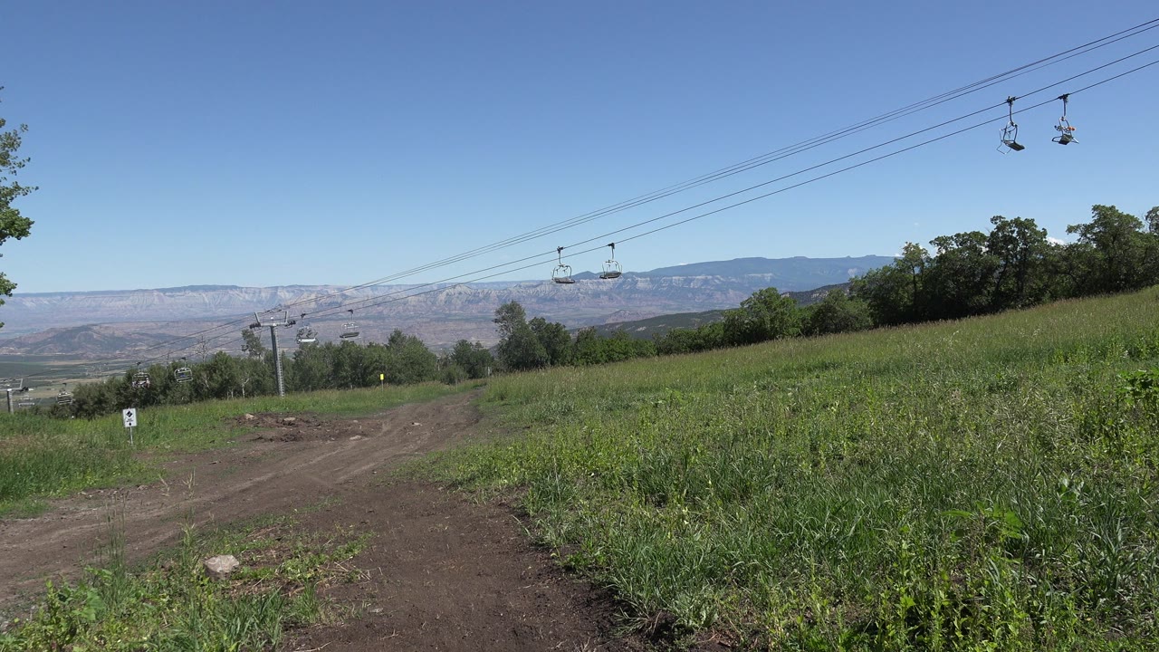 Wide grassy hillside with dirt path, chairlift cables, trees, and distant mountains.