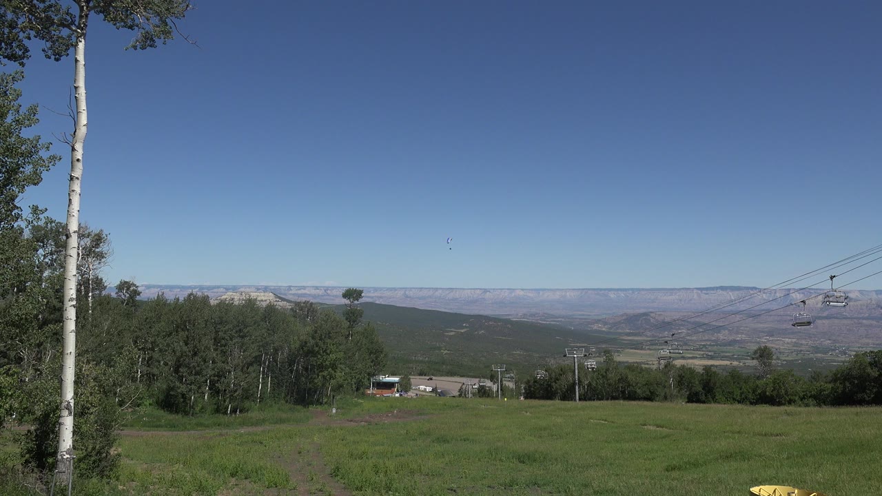 Wide Mesa, Colorado meadow view with green grass in front, pale tree trunks at left, distant mesas on the horizon, chairlift cables at right, and no visible people.