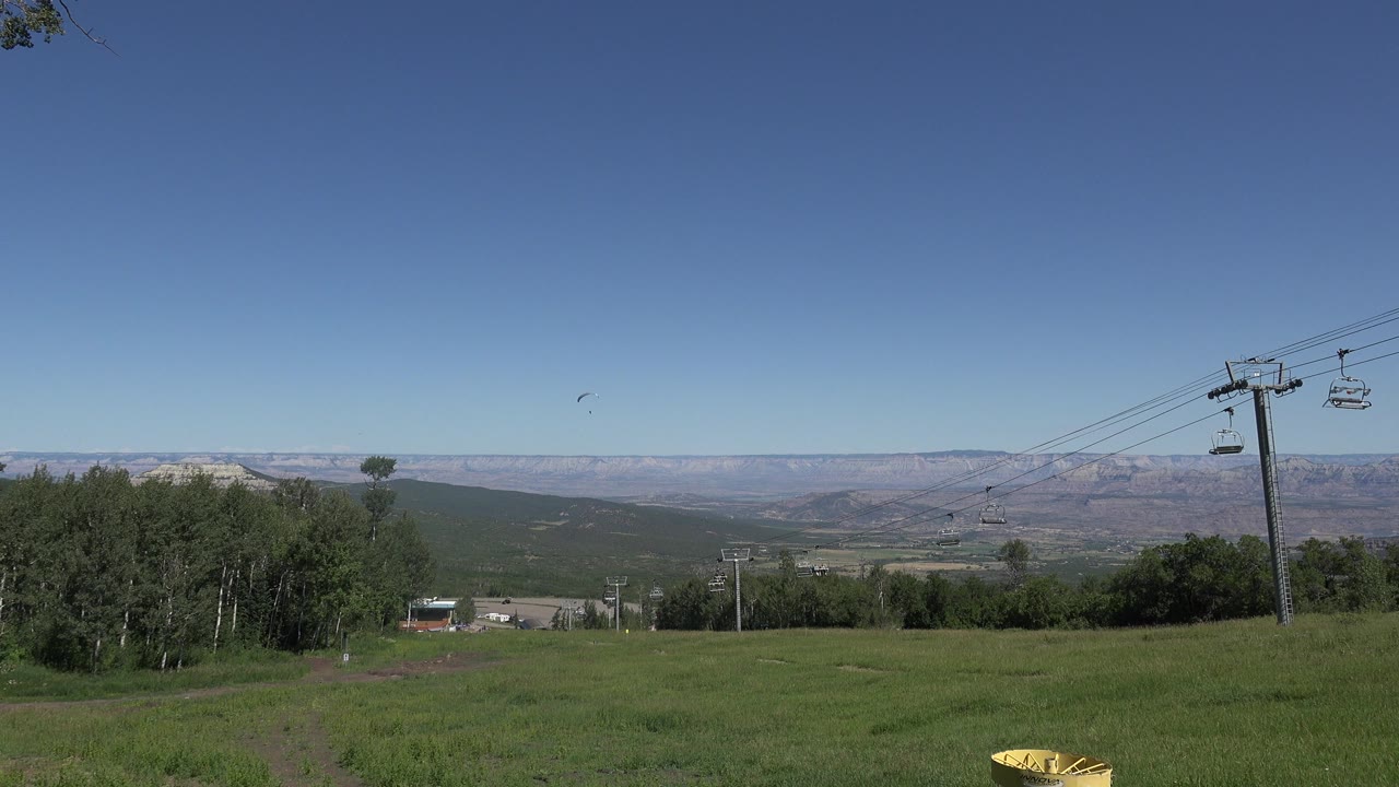 Wide grassy slope with lift cables and distant mesas under blue sky.