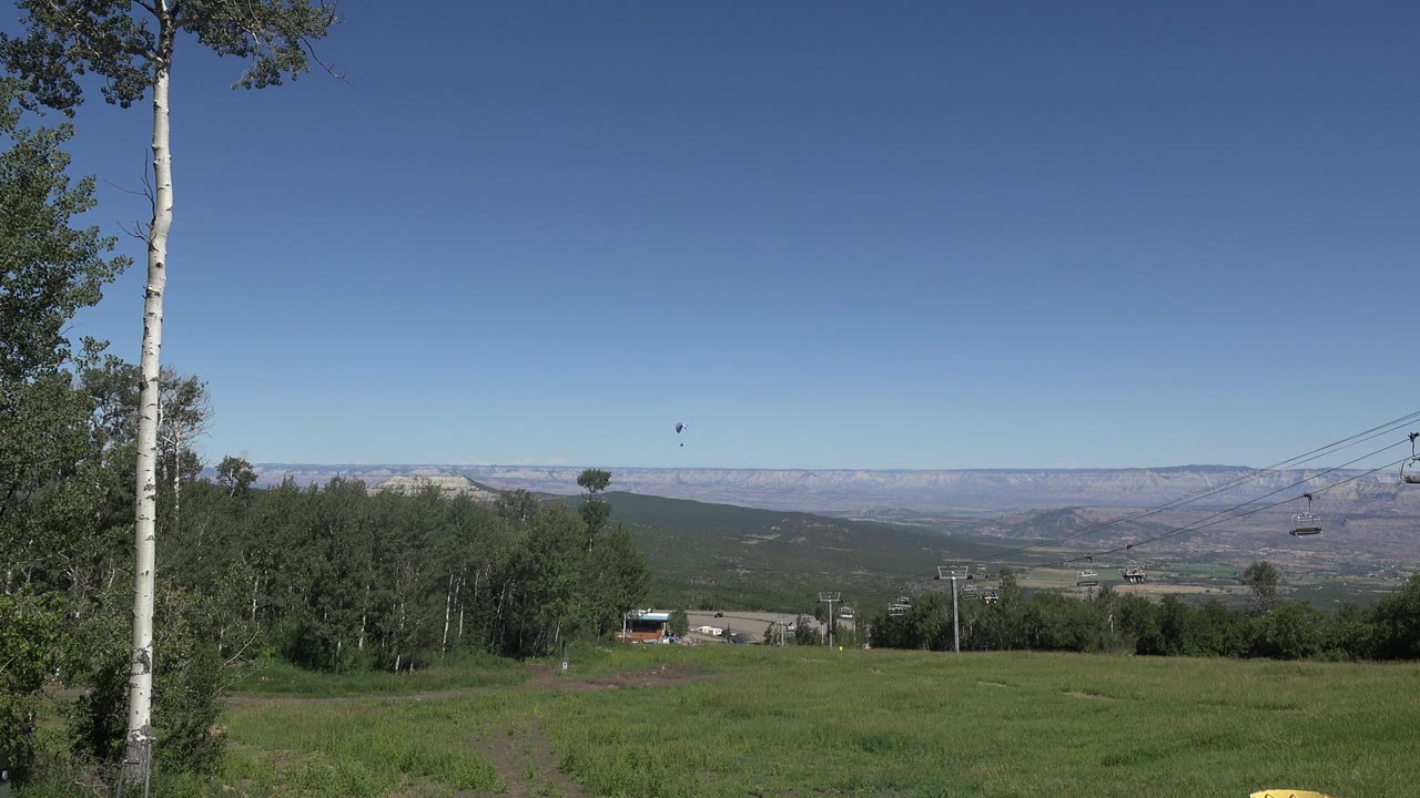 Opening frame of Mesa, Colorado countryside with a green field, white-barked tree at left, distant ridges, chairlift cables at right, a small object in the sky, and no visible people.