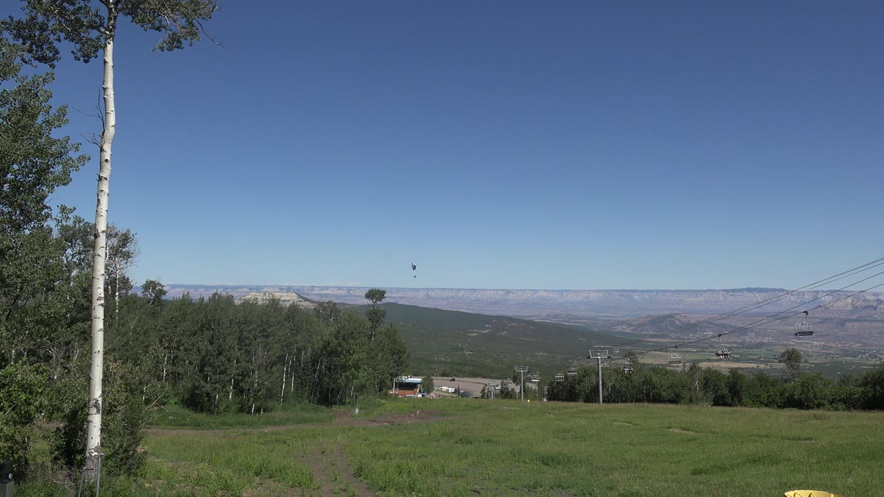 Rural field with trees, distant ridges, lift cables, and a small dark object in the sky.