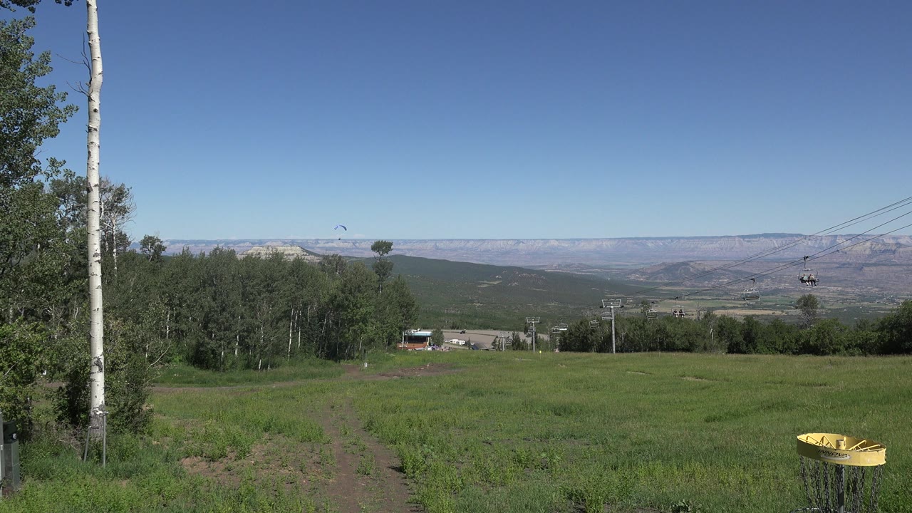 At 0:00 in Mesa, Colorado, a yellow disc golf basket sits at the lower right of a green field with a dirt track, trees, distant ridges, chairlift cables, and no visible people.