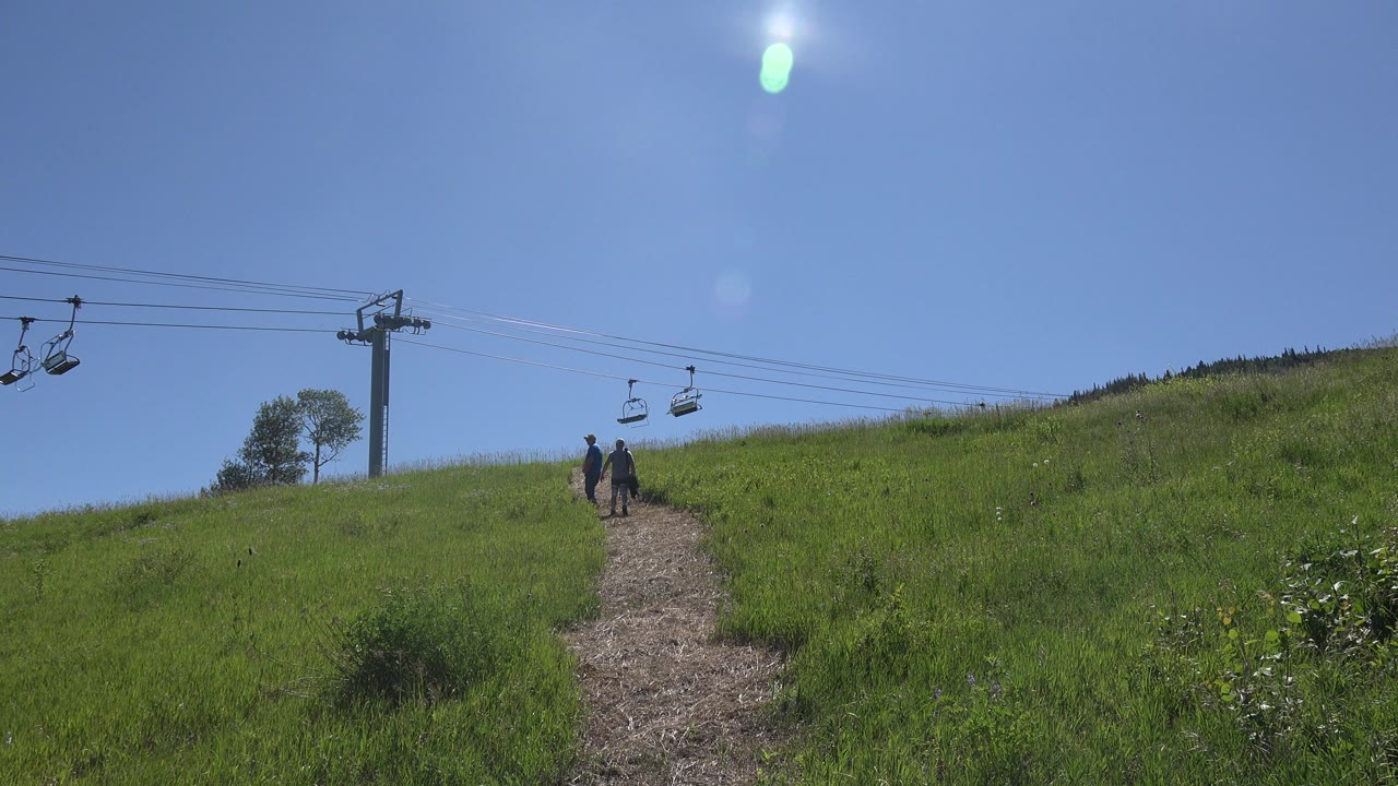 The pair remains near the center of the Mesa hillside path while empty chair lift seats hang on cables above the grassy slope and blue sky.