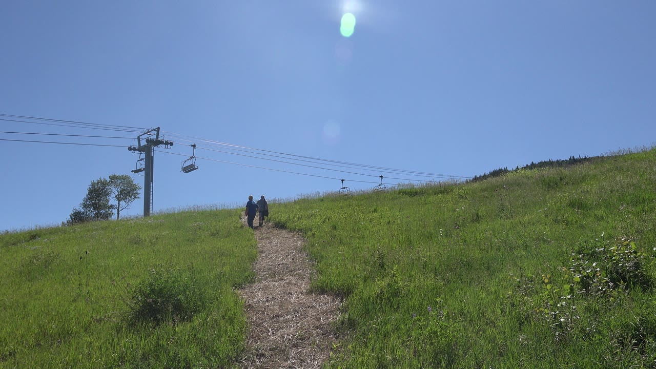 In Mesa, Colorado, distant hikers walk up a narrow hillside trail with green grass on both sides, a ski lift tower at left, and bright lens flare in the blue sky.