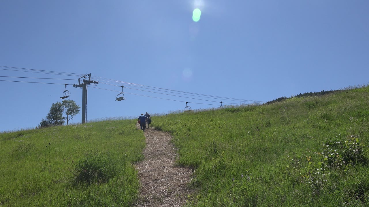 Hikers move farther up a sunlit hillside trail below chairlift chairs.