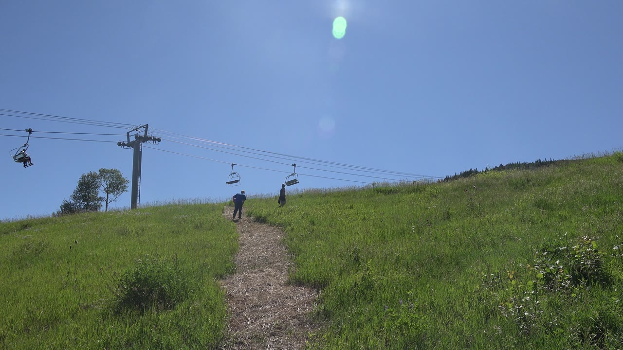 Two people climbing a grassy hillside beneath chairlift cables.