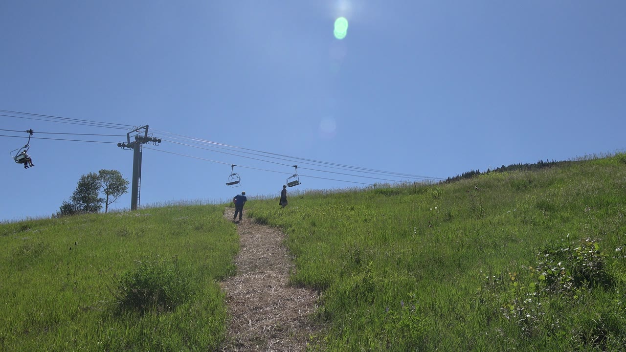 Two small figures continue uphill on a dirt path across a sunlit Mesa, Colorado grass slope, with a lift tower and suspended chairs visible to the left.