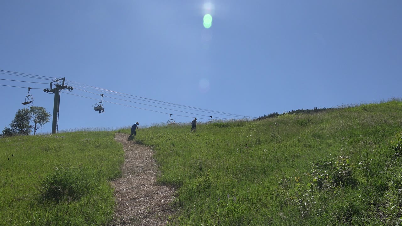 At 0:01 in Mesa, Colorado, the dirt path rises through the grass while one walker stands closer to the path and another remains farther right under the lift line.