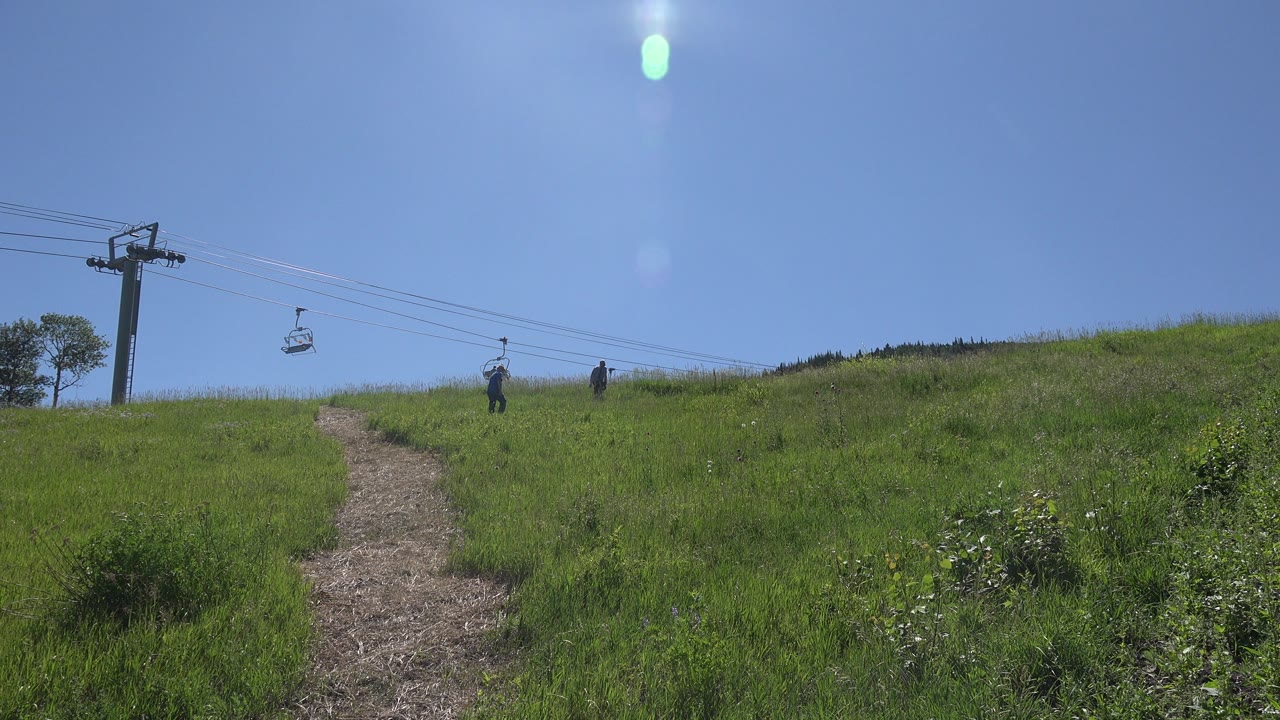 A Mesa, Colorado hillside trail runs up through green grass toward two distant hikers, with ski lift cables and a lift tower under a clear blue sky.
