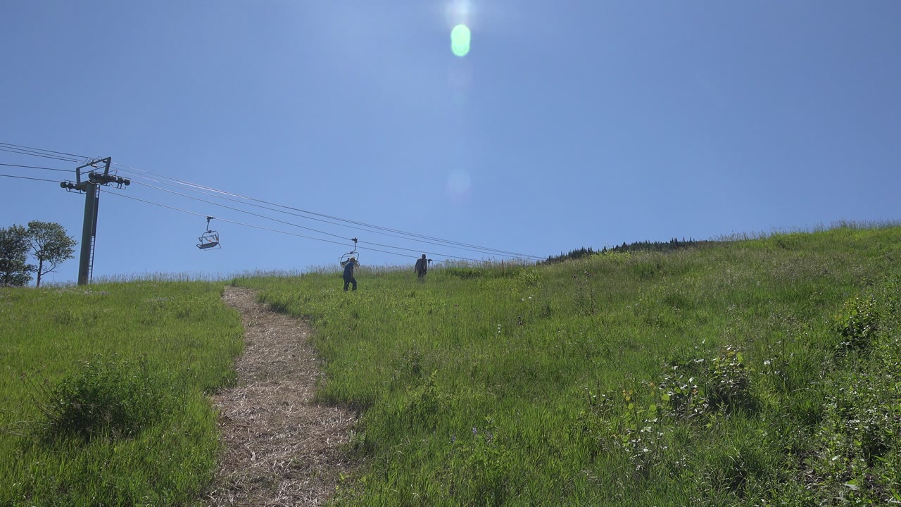 The dirt path remains centered on a green Mesa, Colorado slope as two distant hikers stand near the ridge and chairlift cables cross the sky.