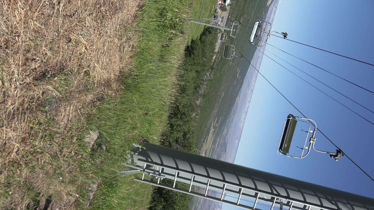 Tilted view of dry grass, green hillside, chairlift cables, and blue sky.