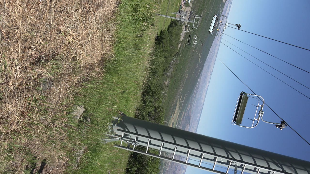 A tilted Mesa, Colorado view holds on dry grass and green slope while chairlift chairs hang from cables against a clear blue sky, with no clearly visible people.