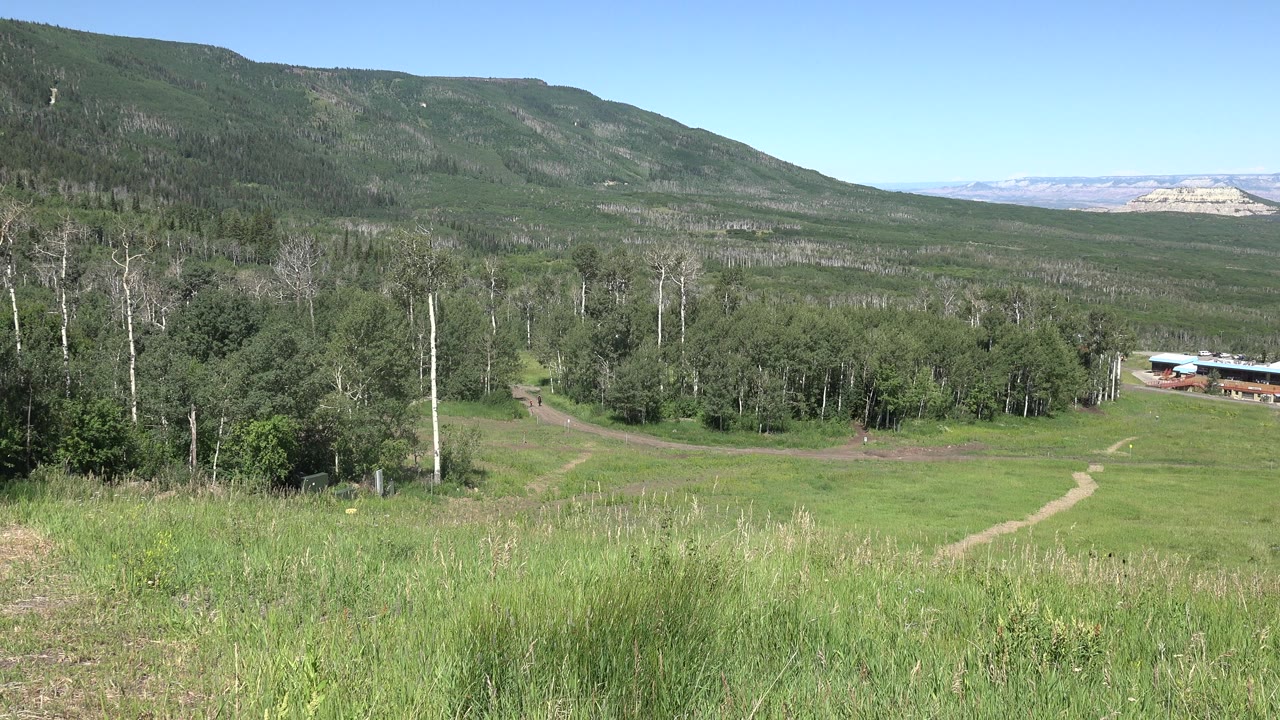 Wide rural landscape with grass, trees, hills, dirt path, and a small building.