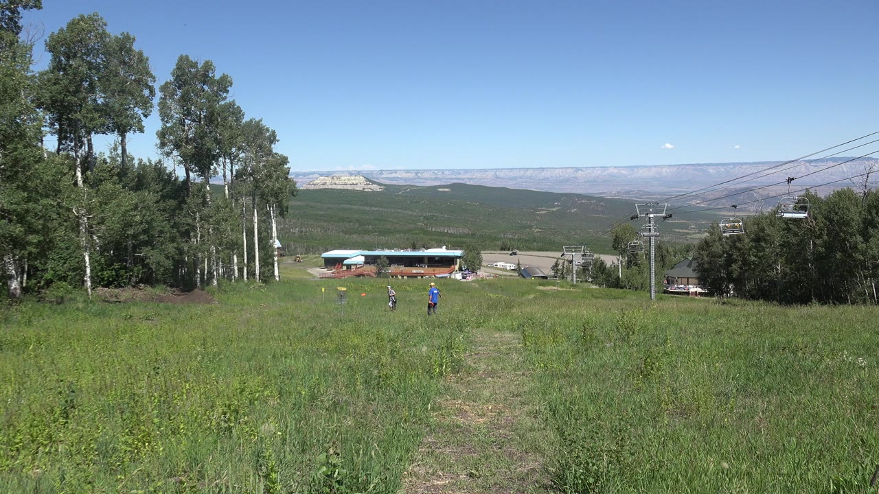 Wide grassy mountain field with two distant people, trees, lift towers, buildings, and blue sky.