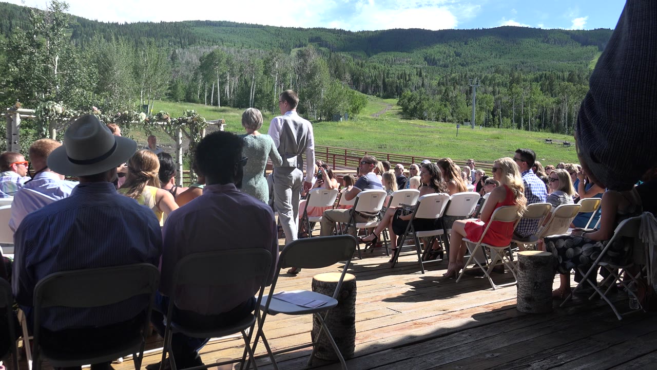Seated wedding guests in Mesa, Colorado face a ceremony arbor on a wooden deck, with green fields and forested hills beyond them.