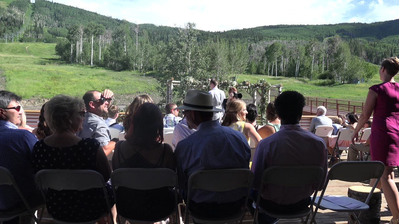 In Mesa, Colorado, seated guests are seen from behind facing a small outdoor ceremony area, with green hills, trees, and bright sky beyond.