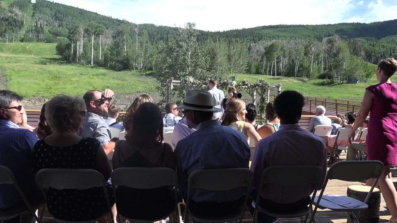 In Mesa, Colorado, rows of guests sit in folding chairs while a woman in a magenta dress stands near the aisle and a floral arch appears ahead.
