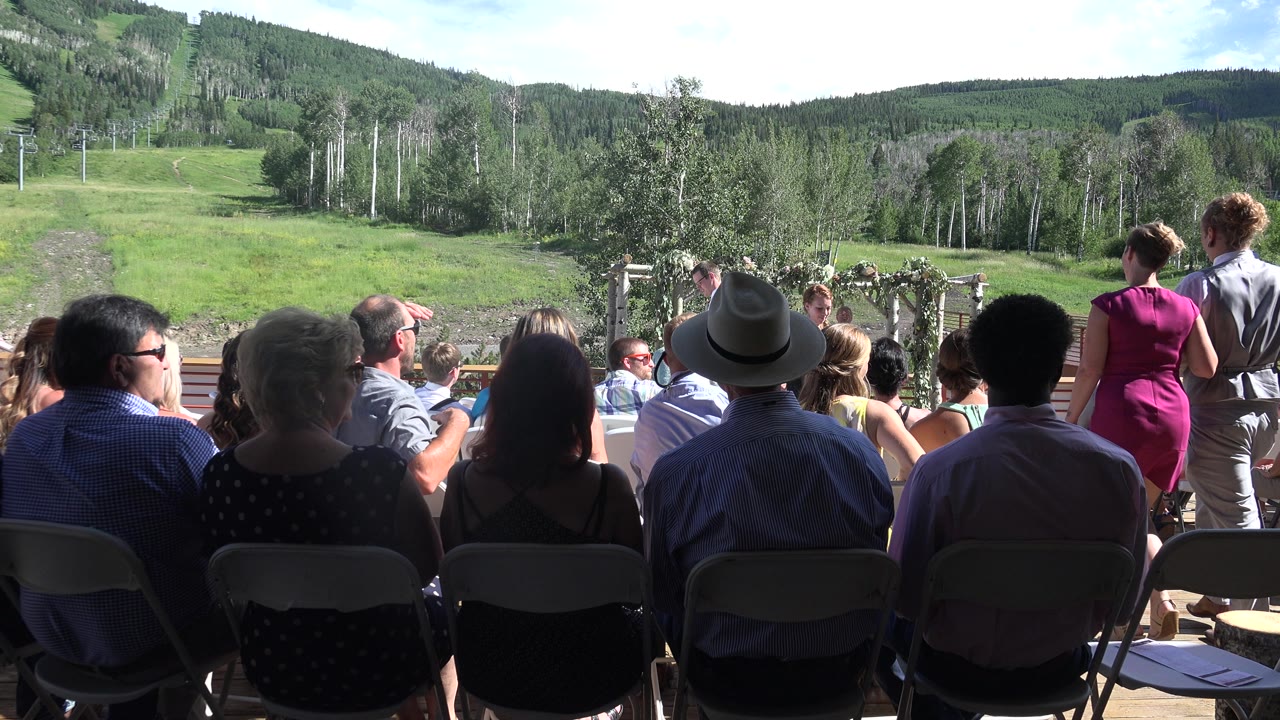 Outdoor audience watches a ceremony below a grassy hillside.