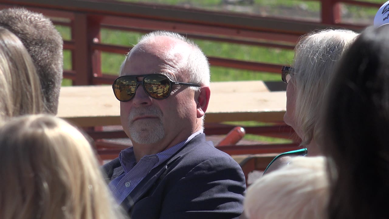 In Mesa, Colorado, the man in sunglasses remains in profile near a blonde woman seen mostly from behind, with other spectators partly blocking the foreground.