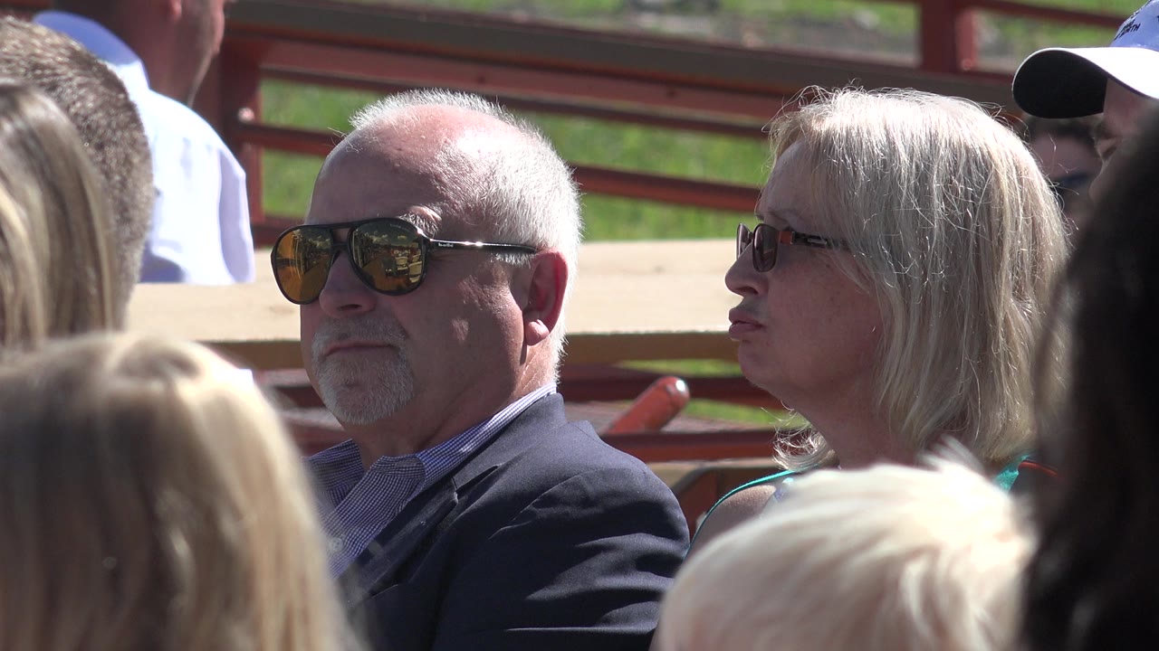 In Mesa, Colorado, a gray-haired man and blonde woman wearing sunglasses sit side by side among blurred attendees in bright sun.