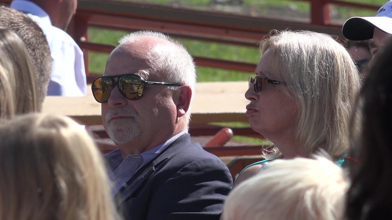 The Mesa crowd cutaway holds on the seated man and woman as foreground heads partly block the lower frame and red railings run behind them.