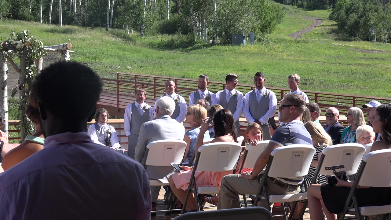 Outdoor gathering with seated guests and men standing beyond white chairs.