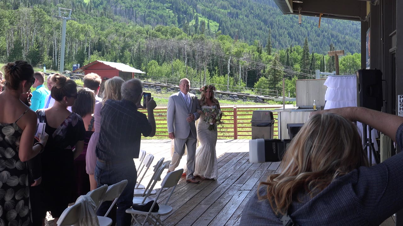 Guests watch a bride and groom standing on a deck with mountains behind them.