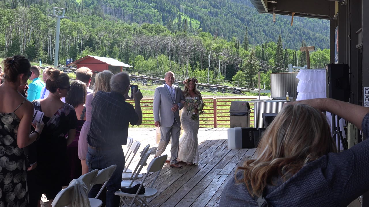 In Mesa, Colorado, the bride and groom pause near the center of the deck while guests gather at left and a photographer points a camera toward them.
